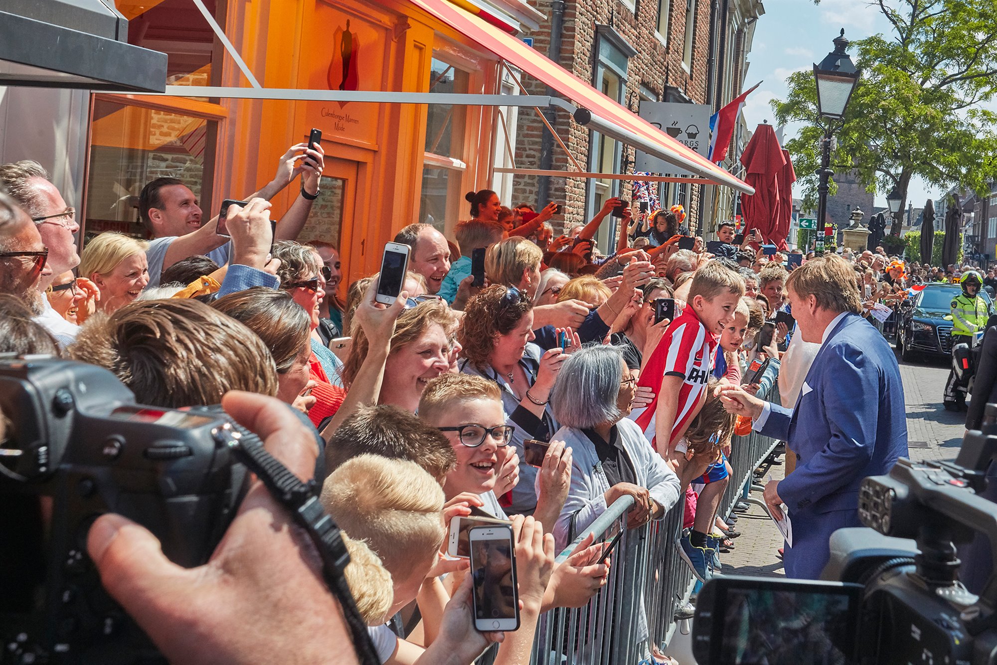 Koning Willem Alexander in gesprek met een van de vele kinderen langs de route. Veel vrolijke gezichten, blije kinderen voor een winkelpand, onder een zonnescherm achter dranghekken langs de looproute van het koninklijk paar. Verder zijn gebouwen, bomen, lantaarnpaal, motoragent, auto en camera's te zien.