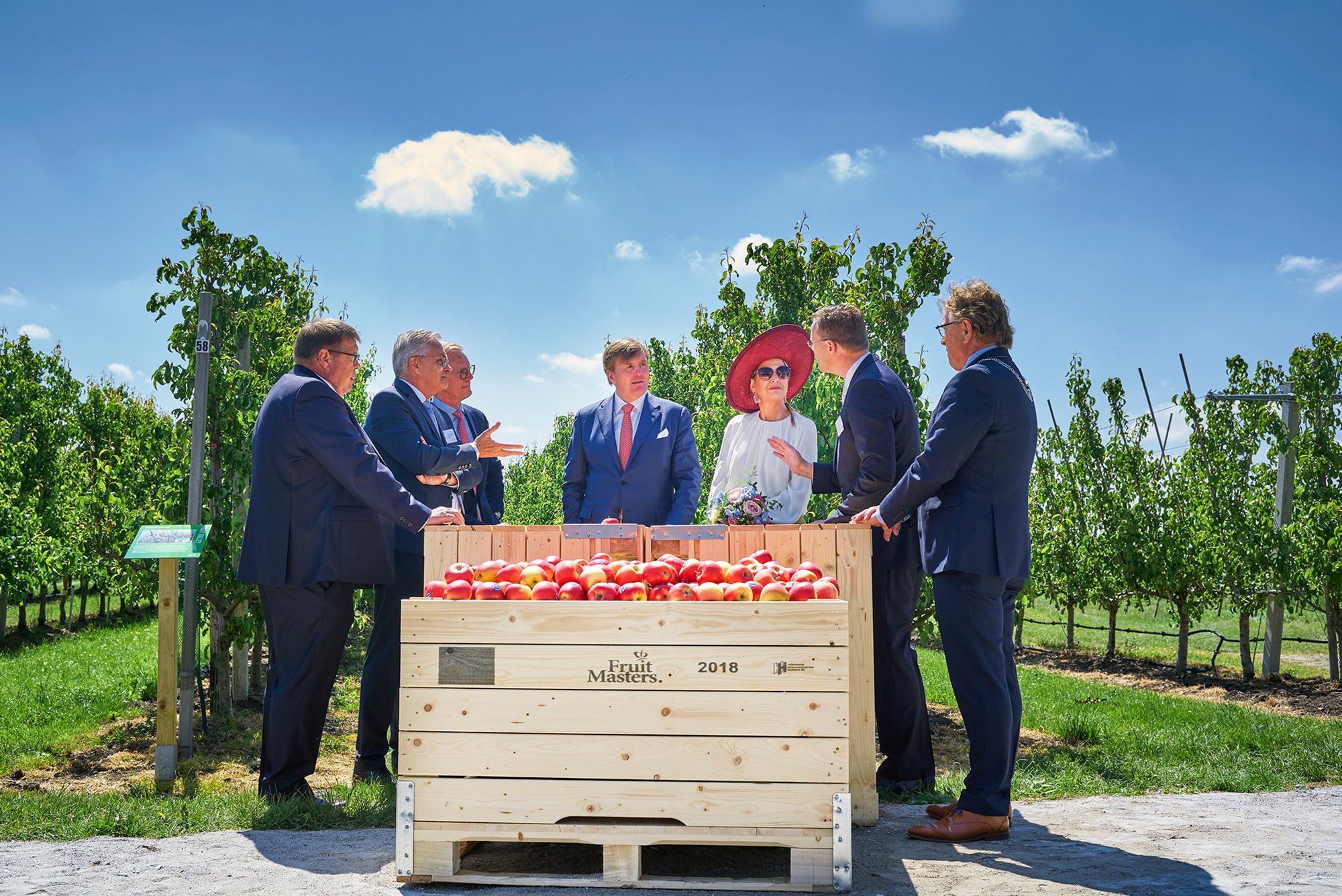 Koning en Koningin in gesprek met bestuurders bij de proeftuin. Op de voorgrond kisten met appels opdruk Fruit Masters 2018, op de achtergrond bomen in de boomgaard en blauwe lucht met kleine witte wolken.