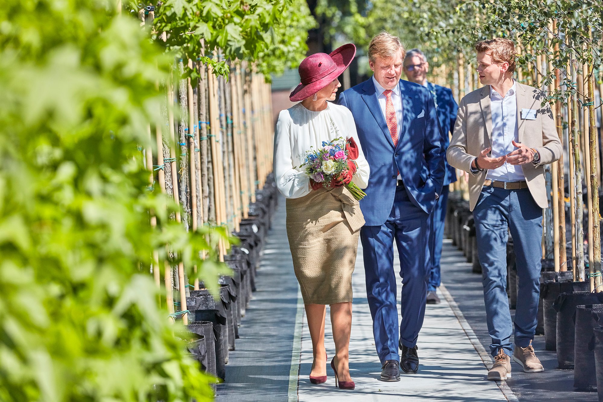 Koning en Koningin luisteren al wandelend tussen de bomenlaan naar een jonge medewerker.