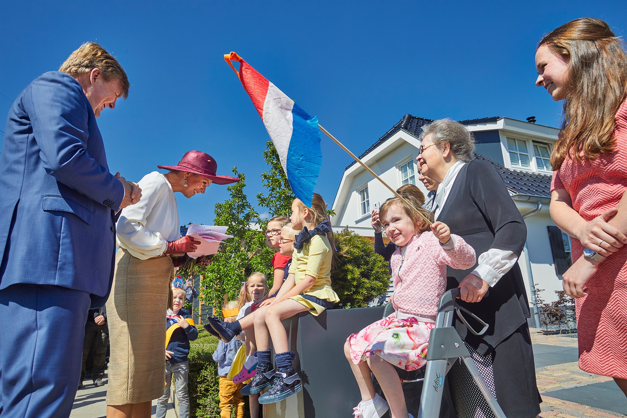 Kinderen langs de route, een meisje zwaait met de Nederlandse vlag, zittend op de rollator van haar oma, Koningin Maxima is in gesprek met enkele jonge kinderen, de Koning luistert mee. Vrolijke gezichte, zonnig tafereel. Op de achtergrond een woonhuis, struiken en een blauwe lucht.