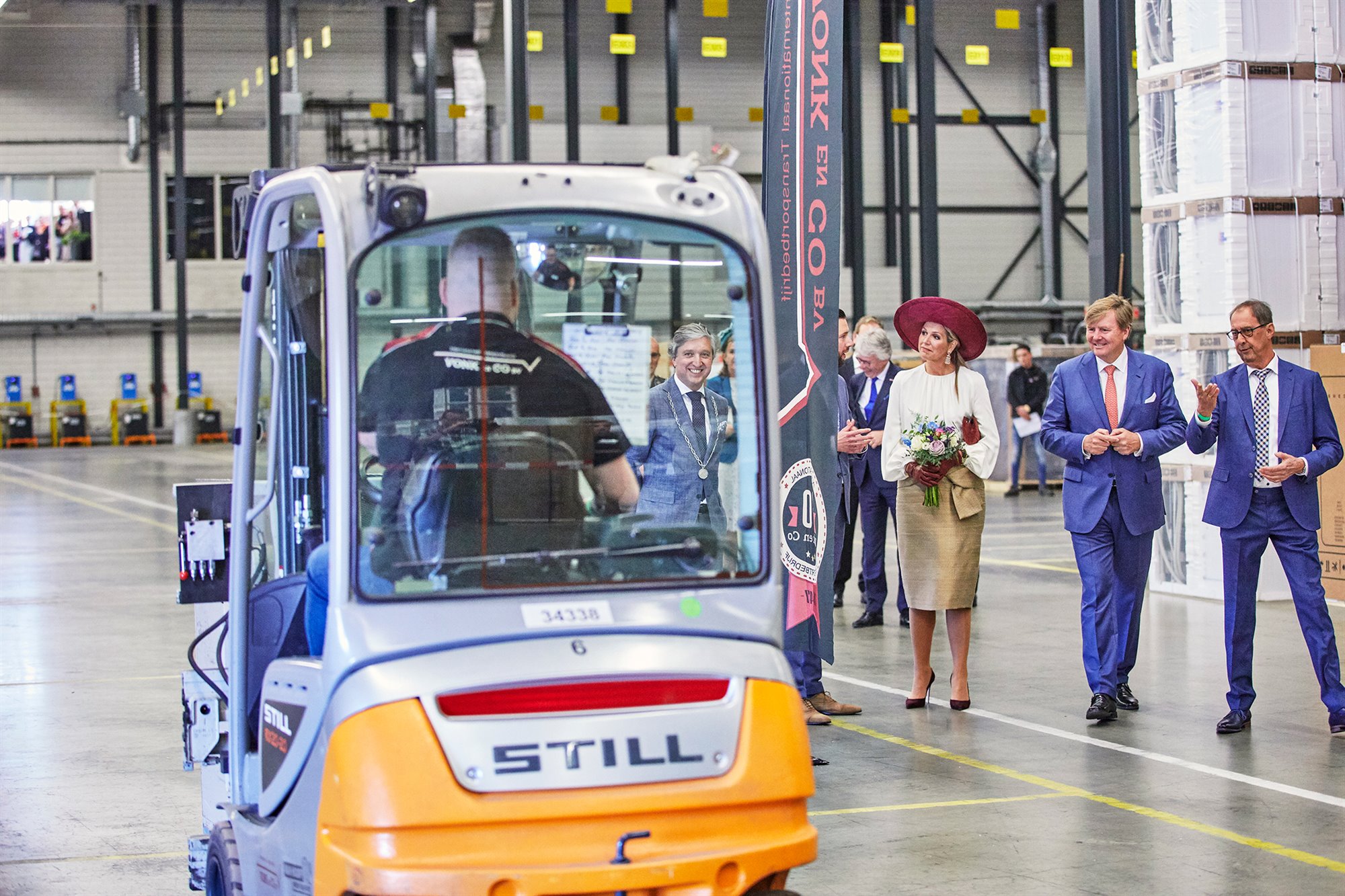 Rondleiding bij Vonk en Co b.v. door de directeur en de Burgemeester in een opslaghal. Op de voorgrond een heftruck die door het magazijn rijdt. Op de achtergrond opgeslagen goederen en betrokkenen en medewerkers.