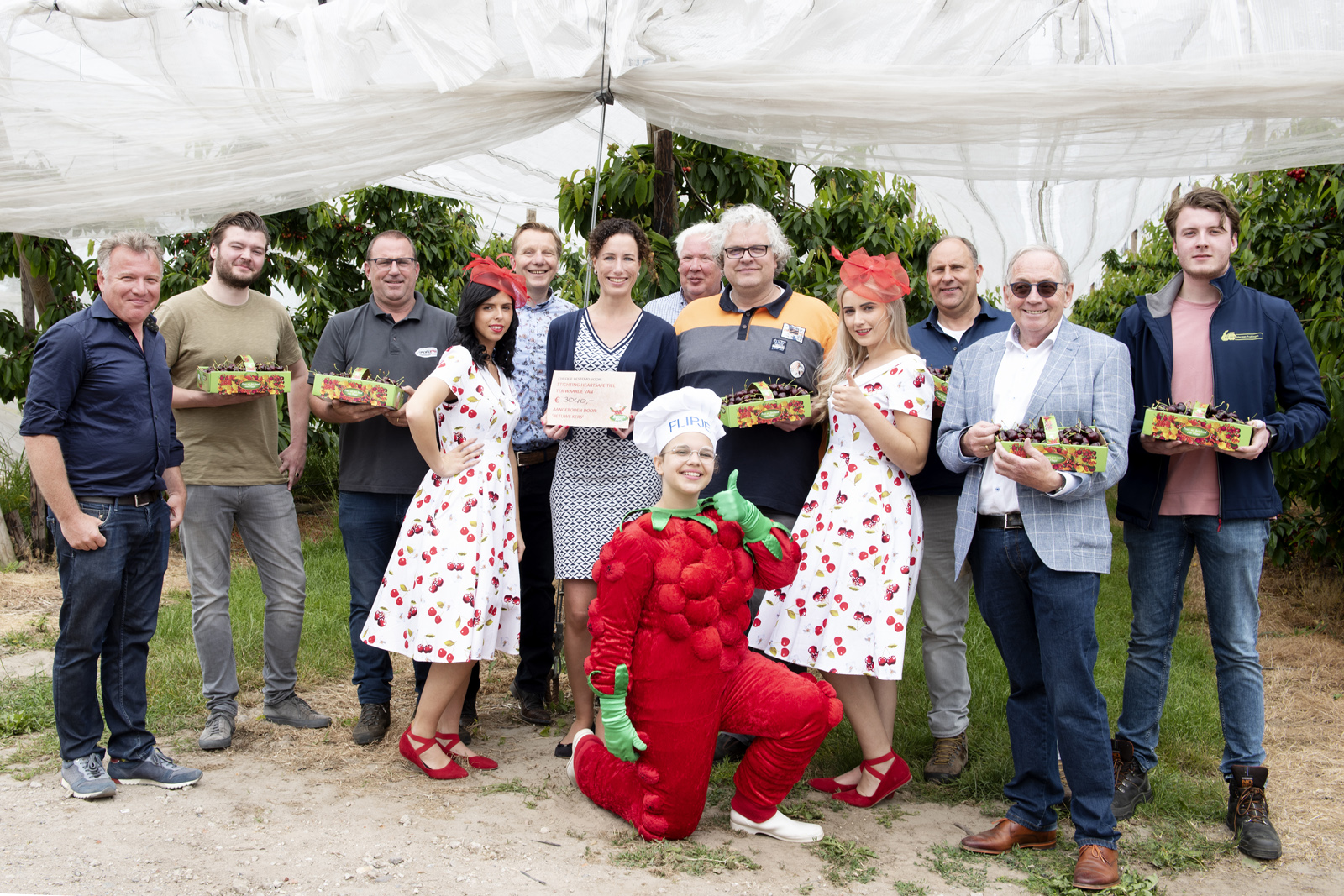 Groep mensen met kistjes kersen, Flipje en de kersenprinsessen voor de kersengaard. Op de achtergrond zijn de kersenbomen met daaroverheen netten gespannen.